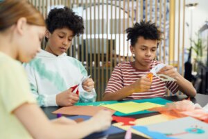 Group of multiethnic children sitting at table and making artworks with colorful paper and scissors in light room