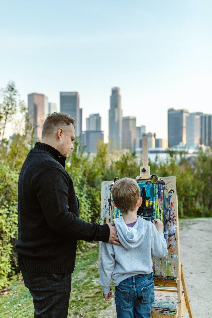 A Man Standing Beside a Child Drawing on a Wooden Easel